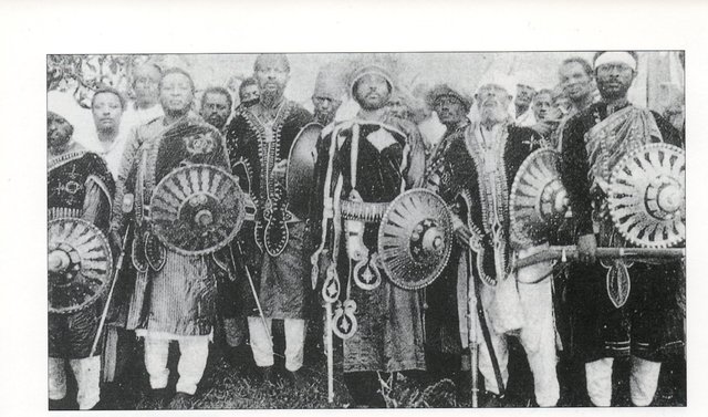Un grupo de importantes guerreros etíopes de las levas feudales observen los escudos con un fino trabajo de filigrana, el del medio posiblemente es un importante Ras de alguna provincia pues lleva el sombrero con piel de león, además de dos con turbantes blancos lo que indica su procedencia musulmana